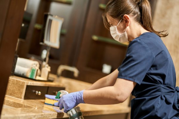 Side view of maid in mask splashing disinfectant on the bathroom surface. Hotel service concept