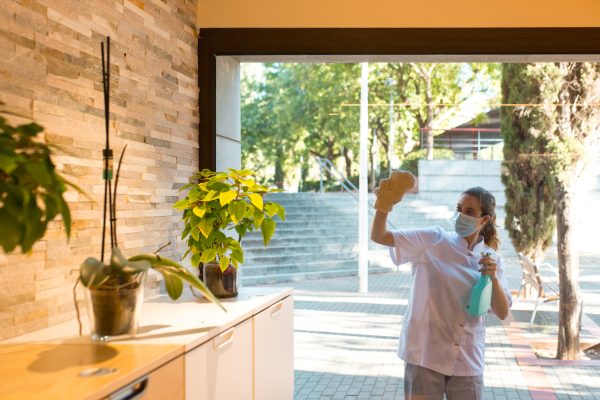 Front view of young woman in uniform and wearing a face mask cleaning windows of spa center from outside.