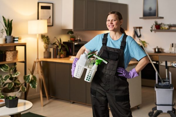 Portrait of woman in uniform with detergent smiling at camera while doing housework in apartment