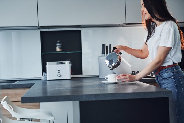 Pouring fresh milk into the glass. Young beautiful brunette in casual clothes indoors in kitchen at daytime.